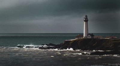 A white lighthouse sits next to a darkened ocean. Dark clouds in the background. A few smaller waves crash.